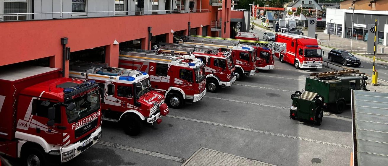 Mehrere rote Feuerwehrfahrzeuge sind vor einer Feuerwache geparkt, mit einem hohen Turm und Bergen im Hintergrund.