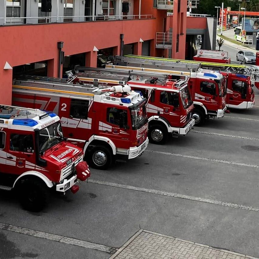 Mehrere rote Feuerwehrfahrzeuge sind vor einer Feuerwache geparkt, mit einem hohen Turm und Bergen im Hintergrund.