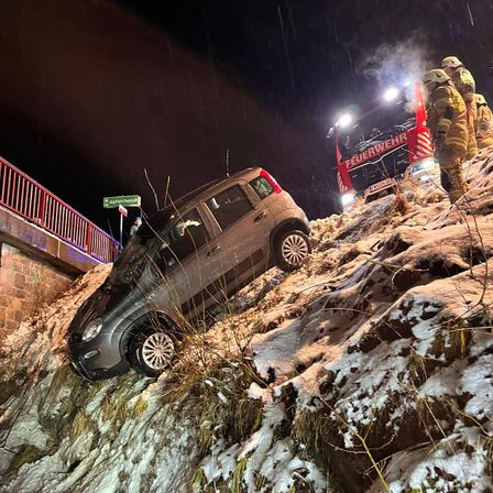 Ein Auto ist auf einem verschneiten Hügel geneigt, mit Feuerwehrleuten in der Nähe. Ein Feuerwehrfahrzeug ist auf der Seite geparkt, und eine Person steht auf dem Zaun. Die Szene ist nachts.
