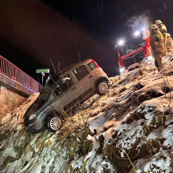 Ein Auto ist auf einem verschneiten Hügel geneigt, mit Feuerwehrleuten in der Nähe. Ein Feuerwehrfahrzeug ist auf der Seite geparkt, und eine Person steht auf dem Zaun. Die Szene ist nachts.