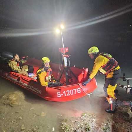 Drei Feuerwehrleute in gelben Helmen und Sicherheitswesten befinden sich in einem roten Boot mit der Aufschrift S-12076. Sie befinden sich nachts in einem Gewässer mit einer montierten Leuchte.