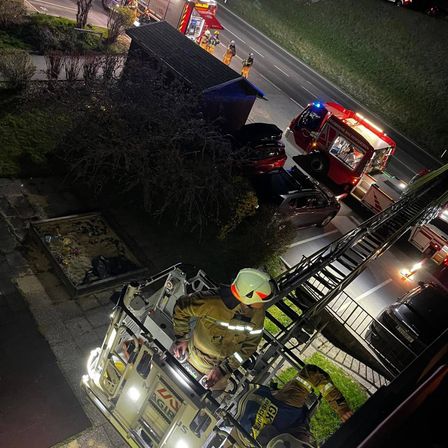 Feuerwehrleute sind am Unfallort mit einem Feuerwehrwagen und einem Auto auf der Straße. Ein Feuerwehrmann in einem Helm klettert eine Leiter.
