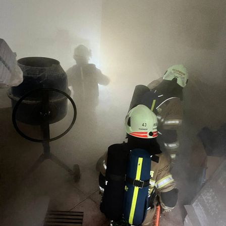Feuerwehrleute in Schutzausrüstung navigieren durch einen rauchgefüllten Raum, wobei einer einen Schlauch trägt. Im Hintergrund ist die Silhouette eines anderen Feuerwehrmanns zu sehen.
