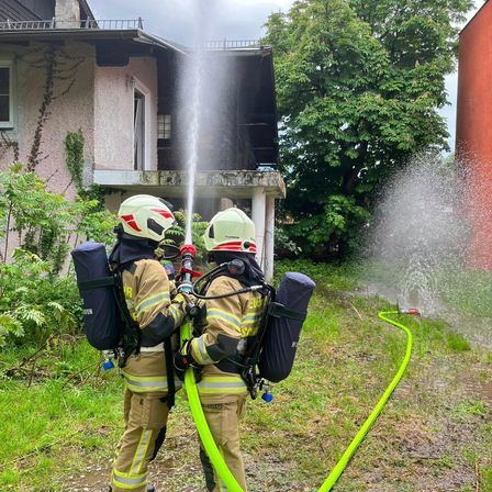 Zwei Feuerwehrleute in voller Ausrüstung spritzen Wasser aus einem Schlauch vor einem Haus mit offenem Fenster.