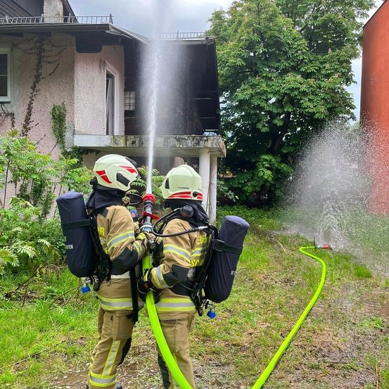 Zwei Feuerwehrleute in voller Ausrüstung spritzen Wasser aus einem Schlauch vor einem Haus mit offenem Fenster.