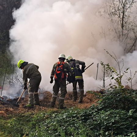 Drei Feuerwehrleute mit Schutzausrüstung arbeiten an einem Waldbrand. Sie sind von Rauch umgeben und benutzen Werkzeuge, um das Feuer zu löschen.