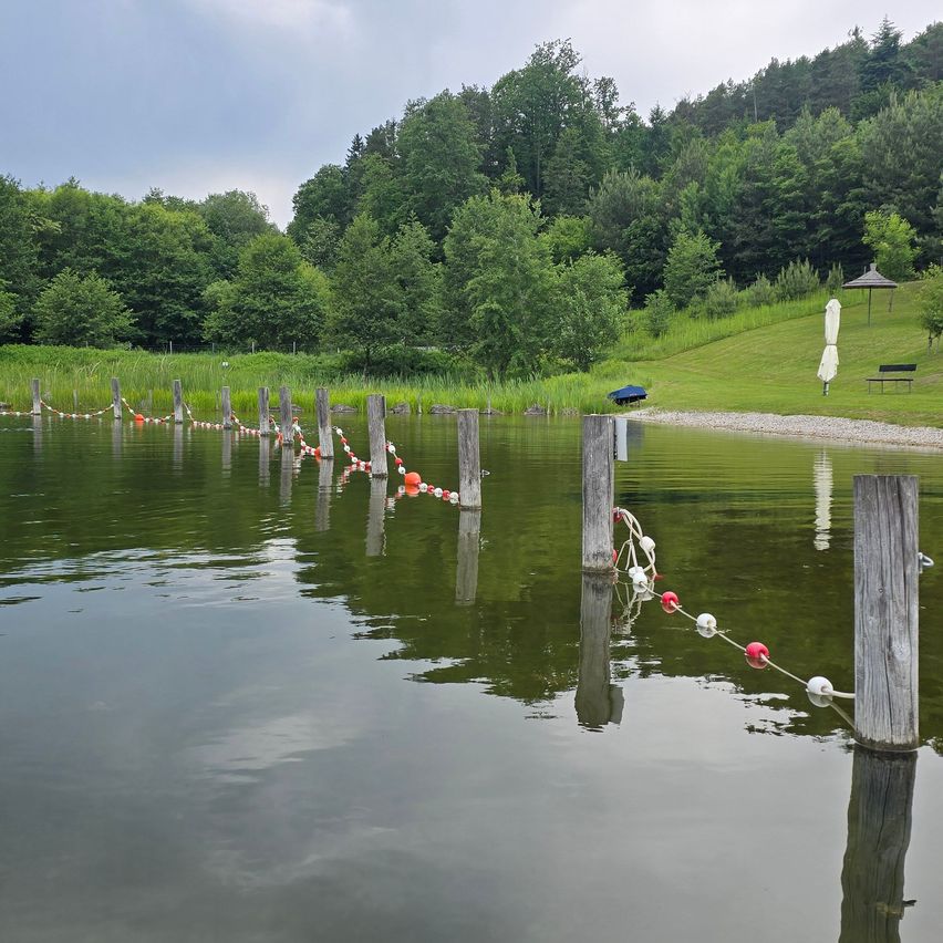 Ein ruhiger See mit einer Reihe von Holzpfählen, verbunden durch eine Linie roter und weißer Bojen, umgeben von üppigem Grün und einem entfernten Schirm auf einem Grashügel.