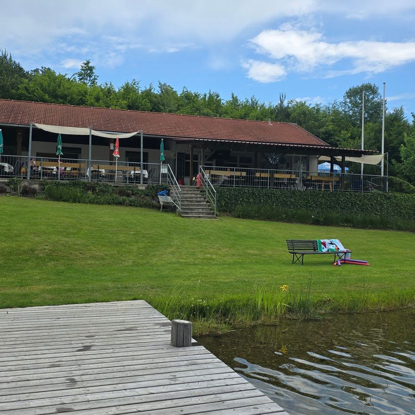 Ein Gebäude am Seeufer mit Terrasse, Treppen und Tischen mit geschlossenen Schirmen. Das Gebäude hat ein rotes Dach und ist von grünem Gras und Bäumen umgeben.