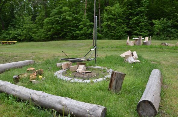 Ein Grasfeld mit Baumstämmen, einem Feuerstellen und einer Metallstruktur. Bäume und Bänke sind im Hintergrund.