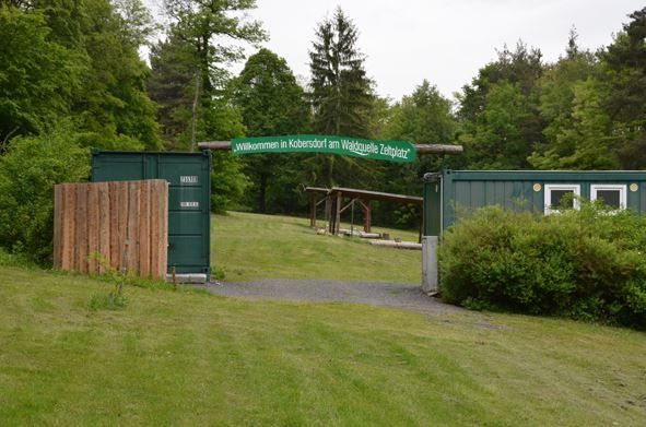 Grüne Container markieren den Eingang zu einem Rasengebiet in Kobersdorf, Österreich, mit einem Holzzaun und einem grünen Schild, das Besucher auf dem Waldquelle Zeltplatz willkommen heißt.
