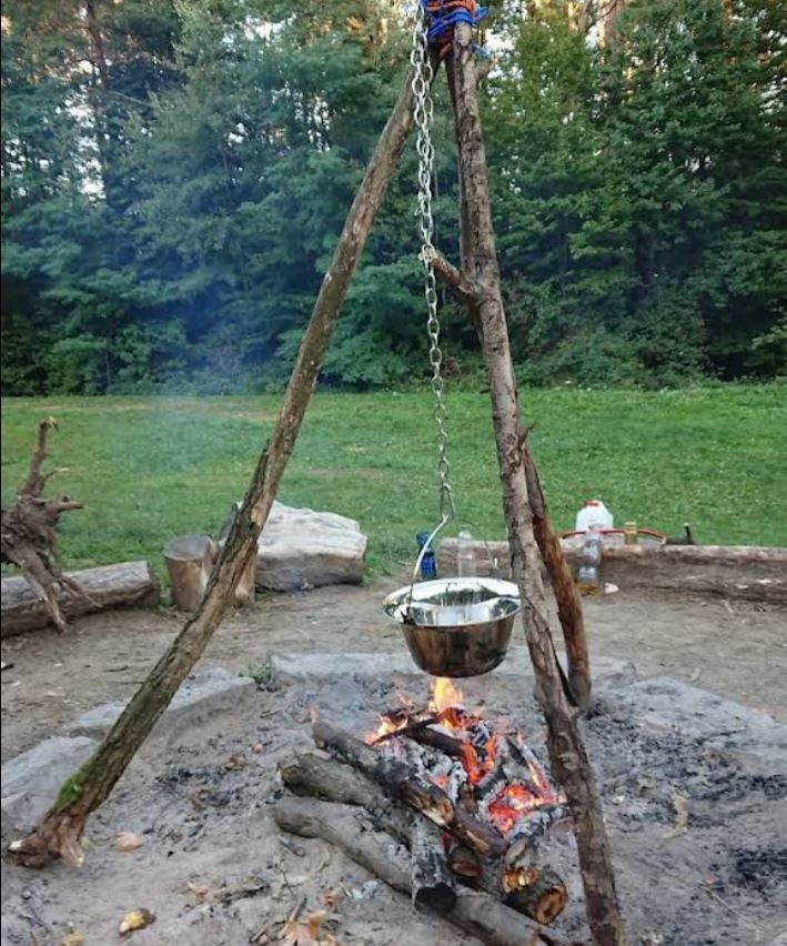 Ein Metalltopf hängt über einem Feuerstellen aus Holzstangen, mit einem Grasfeld und Bäumen im Hintergrund.