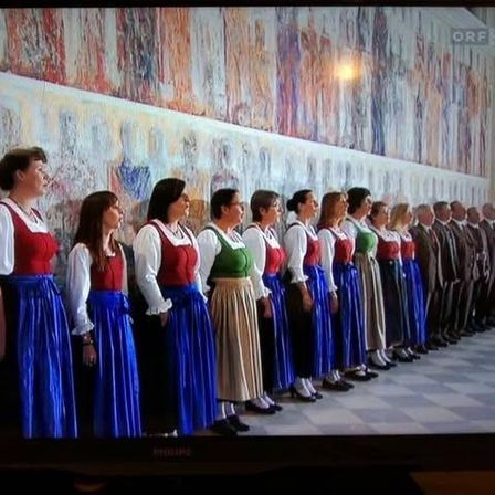 Eine Gruppe von Frauen und Männern in traditioneller Kleidung steht in einer Reihe, der Wand mit Wandgemälden zugewandt und scheint zu singen.