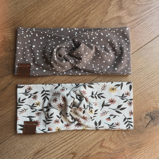 Zwei Stirnbänder auf einer Holzoberfläche. Eines hat ein braunes Punktmuster und das andere ein Blumendesign. Beide haben braune Etiketten.