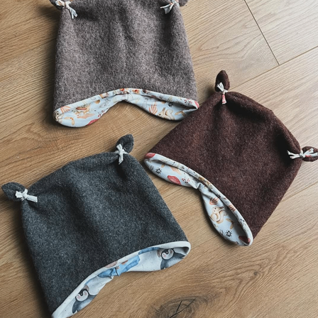 Drei gestrickte Mützen liegen auf einem Holzboden. Sie sind grau, braun und dunkelbraun und jeweils mit dekorativen Elementen und einem kleinen Anhängeband versehen.