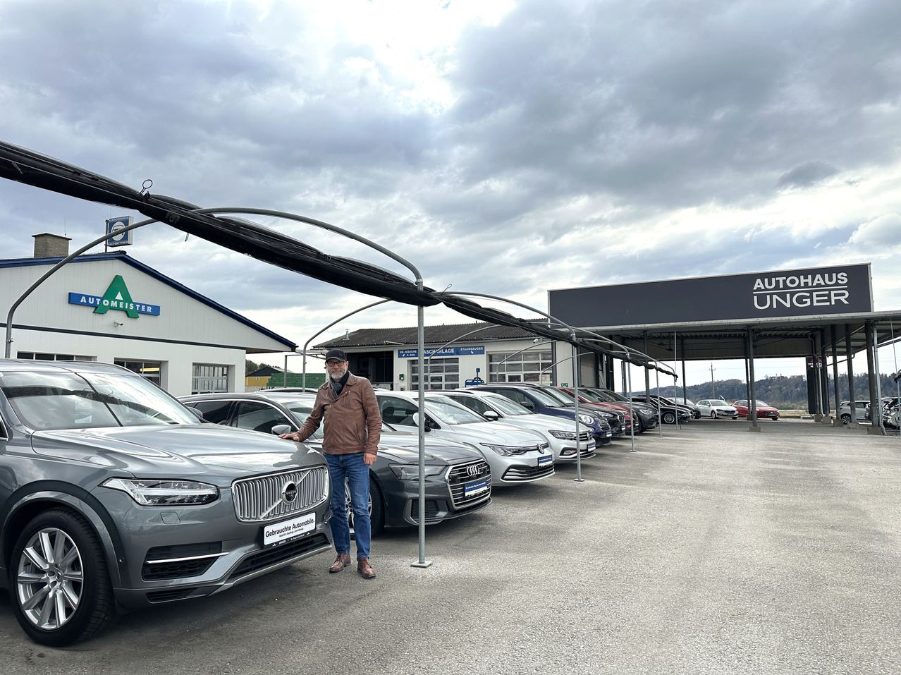 Ein Mann steht neben einem grauen Volvo-Auto, trägt eine braune Jacke und Jeans, mit mehreren Autos, die unter einem Baldachin im Hintergrund geparkt sind.