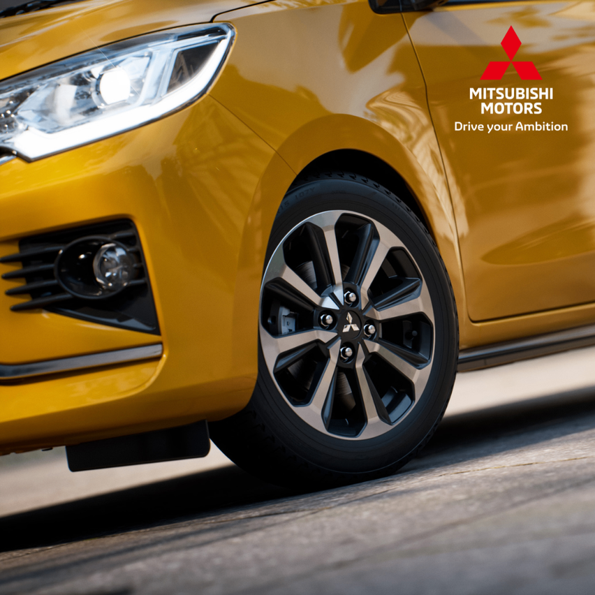 Eine Nahaufnahme eines gelben Mitsubishi-Autos mit dem Slogan 'Fahren Sie Ihre Ambition'. Das Auto zeigt einen Scheinwerfer, ein Logo und einen Reifen mit silbernen Felgen.