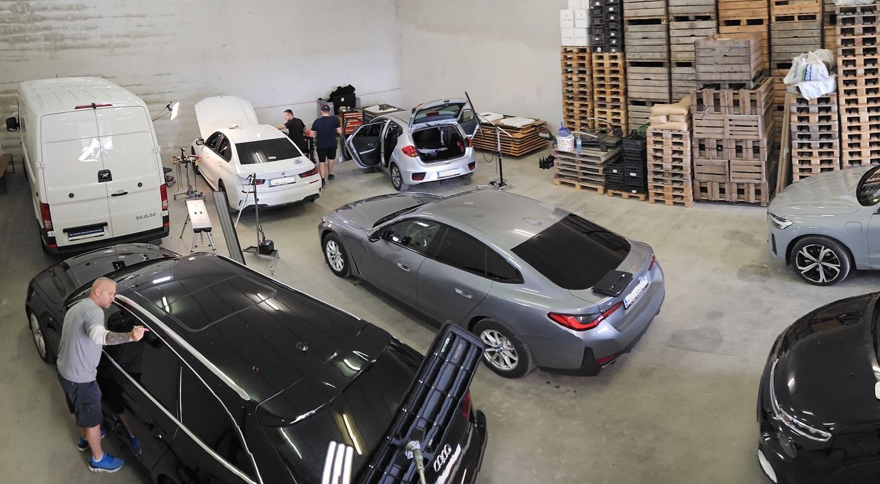 Eine Autowerkstatt mit drei Autos. Zwei Autos haben ihre Motorhauben hochgeklappt und eines hat seinen Kofferraum geöffnet. Arbeiter stehen um die Autos herum. An der Wand sind Holzpaletten gestapelt.