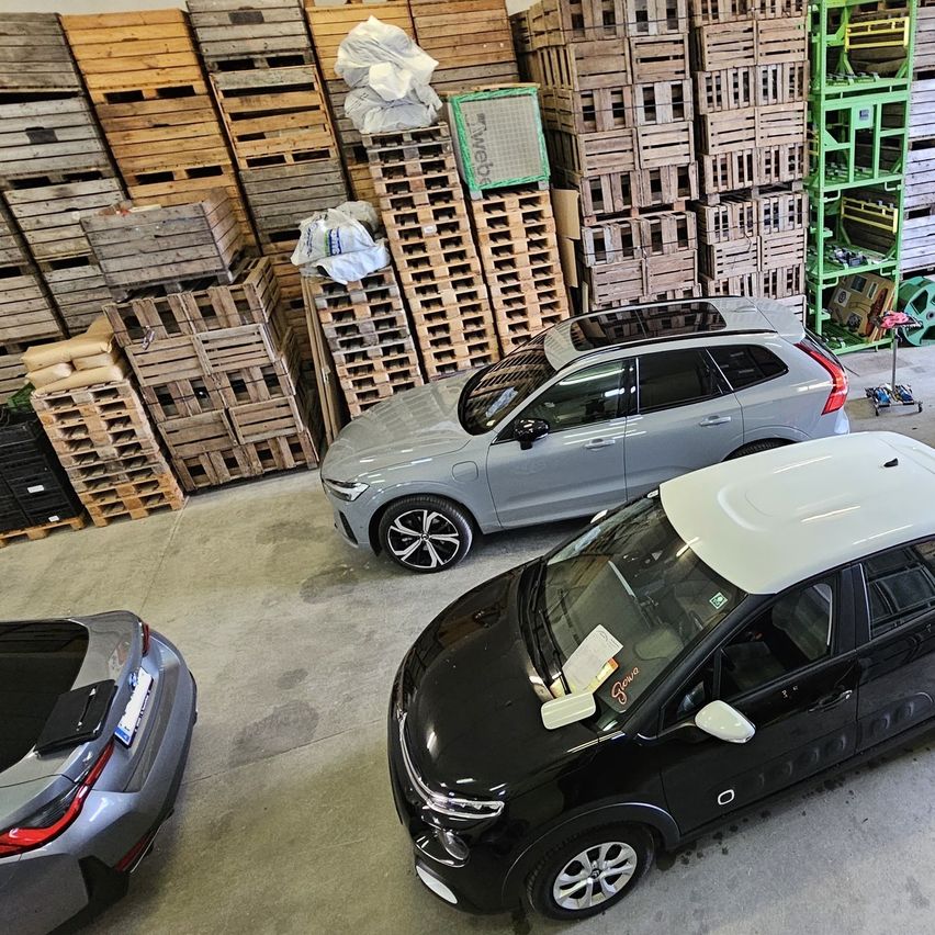 Eine Draufsicht auf ein Lagerhaus mit mehreren gestapelten Holzpaletten, einem grauen Auto, einem schwarzen Auto und einem silbernen Auto, das davor geparkt ist.
