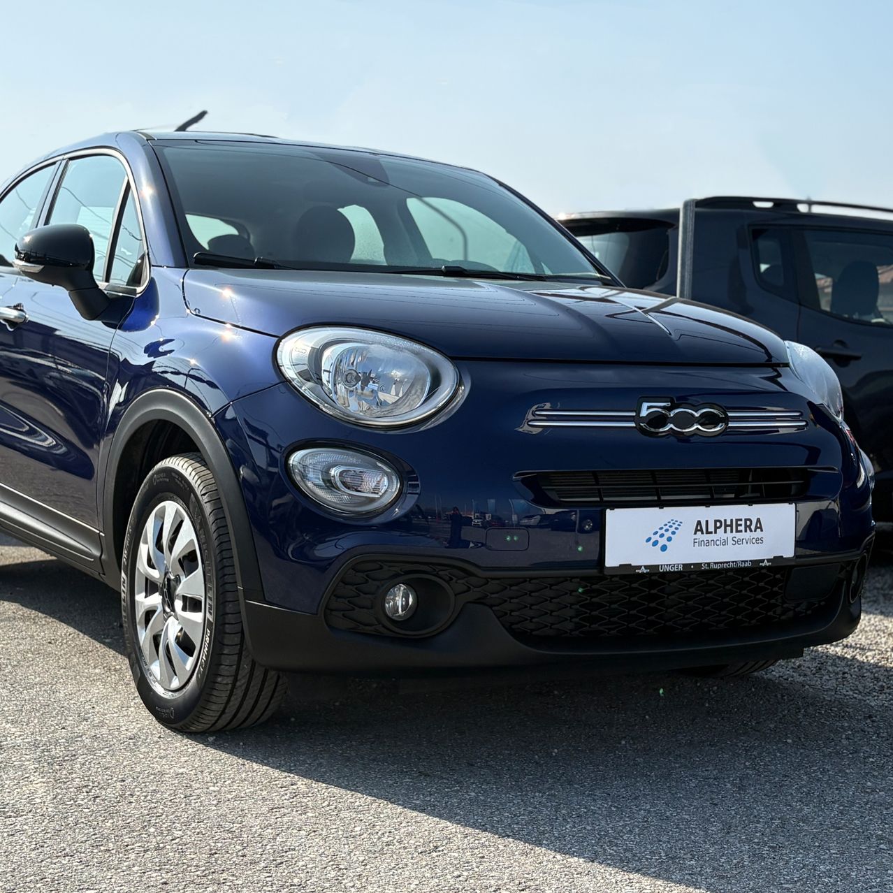 Ein blauer Fiat 500 mit einem Nummernschild, das 'Unger' und das Logo von Alfara Financial Services liest, geparkt auf einer Kiesoberfläche.