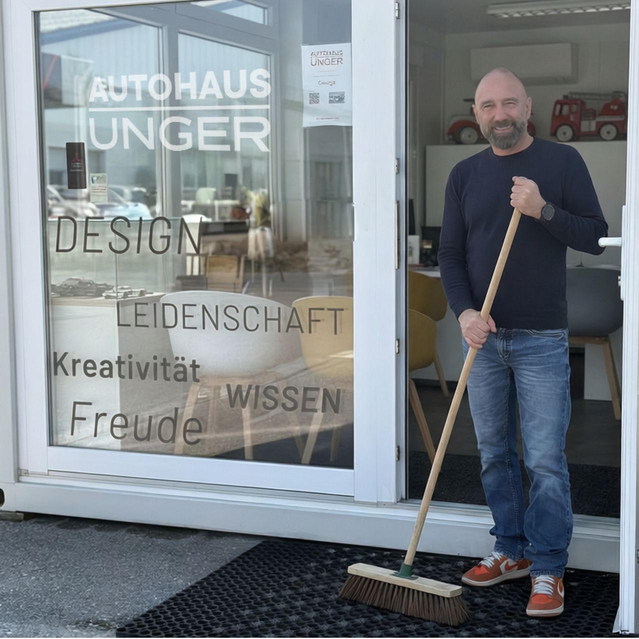 Ein Mann steht vor einer Autohaus Unger-Filiale, hält einen Besen und lächelt. Die Glastür hat Wörter wie Design, Leidenschaft und Freude.