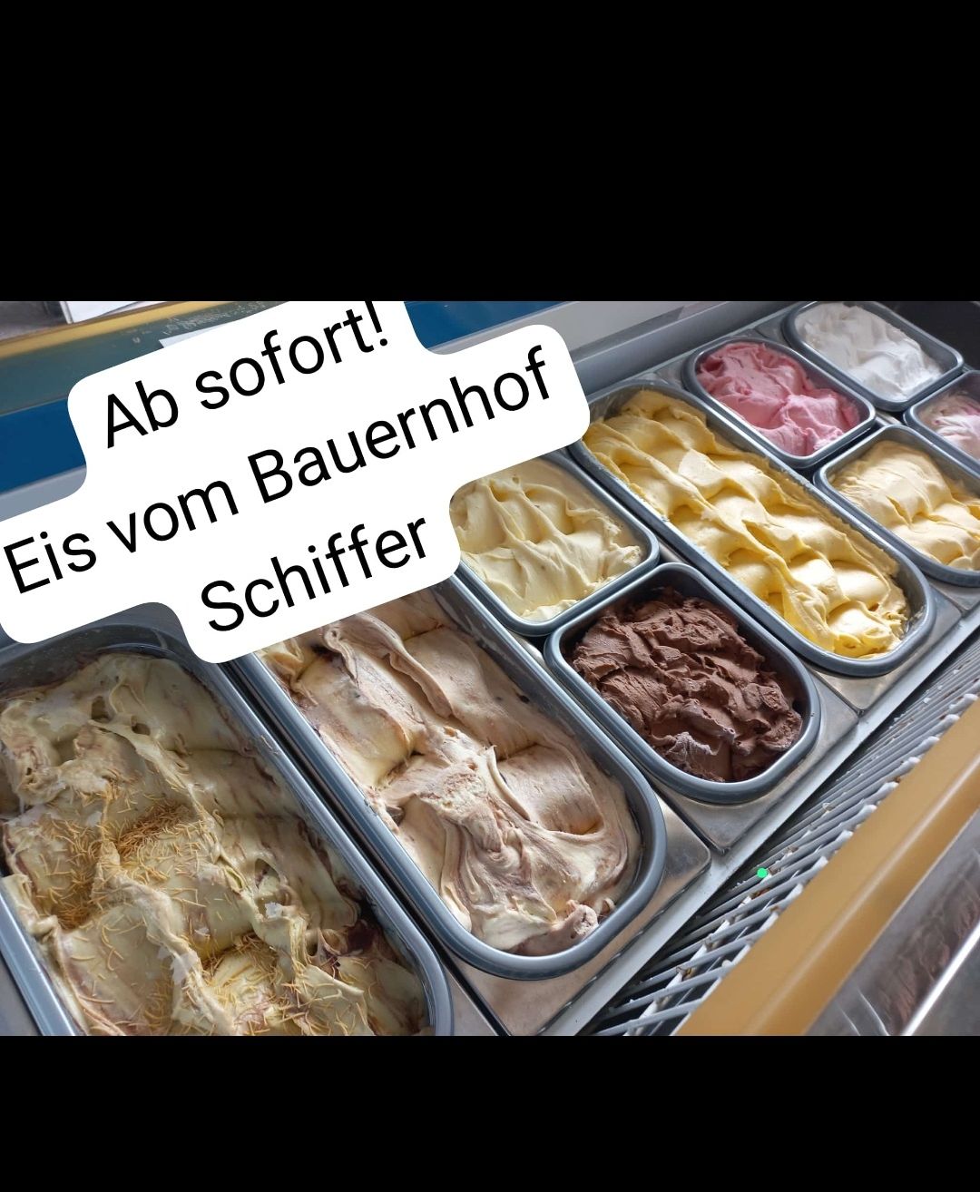 Eine Auswahl an Eissorten in einer gekühlten Vitrine, mit Schildern auf Deutsch, die 'Ab sofort!' und 'Eis vom Bauernhof Schiffer' werben.
