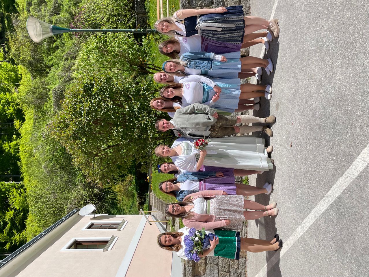 A group of people, mostly women, are smiling and posing for a photo on a road. They are standing in rows, and some are holding flowers. There is a building with windows and a streetlight on the left.