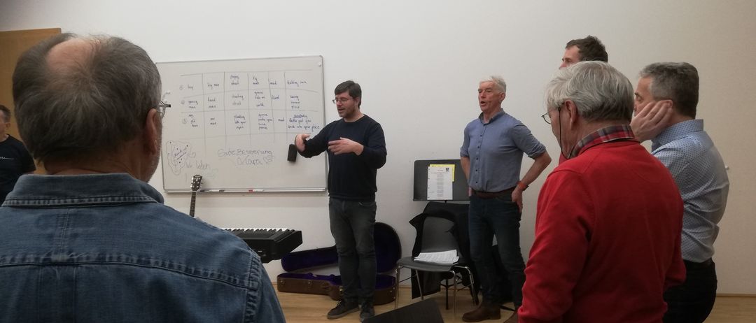 Ein Mann in einem schwarzen Shirt gestikuliert und spricht mit einem anderen Mann in einem blauen Hemd in einem Klassenzimmer. Im Hintergrund sind eine Tafel, Stühle und Musikinstrumente zu sehen.