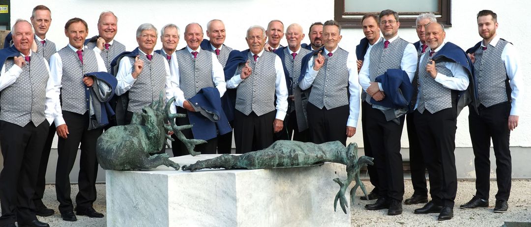 Eine Gruppe älterer Männer, gekleidet in graue Westen und schwarze Hosen, posiert mit einer Skulptur vor einem Gebäude mit einem Schild, das 'KUCHL' steht.