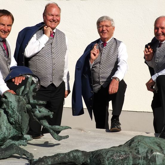 Vier ältere Männer posieren für ein Foto mit einer Bronzeskulptur. Sie tragen formelle Kleidung und stehen vor einer weißen Wand.