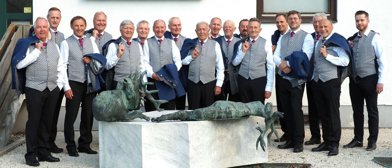 Eine Gruppe von Männern, die Westen und Fliegen tragen, posiert für ein Foto vor einem Schild, das Kuchl steht. Sie stehen hinter einer weißen Skulptur mit einem Hirsch und einer Frauenfigur.