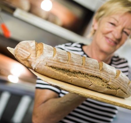 Eine Frau hält ein Brot auf einem Holzbrett in einer Küche mit einem Ofen im Hintergrund.
