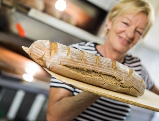 Eine Frau hält ein Brot auf einem Holzbrett in einer Küche mit einem Ofen im Hintergrund.