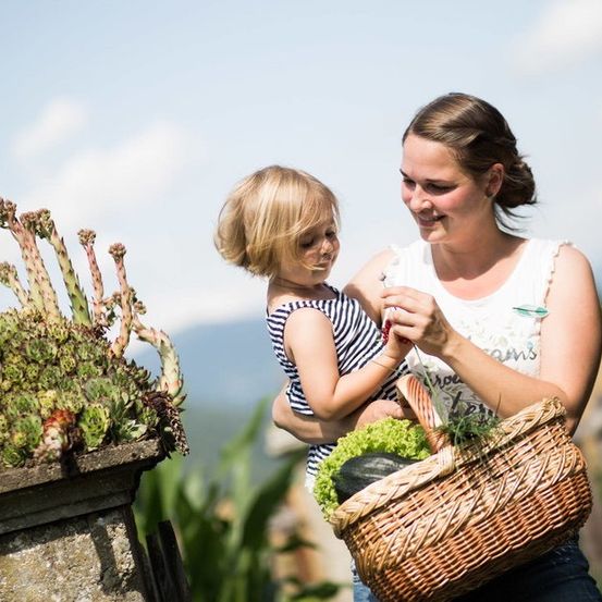 Eine Frau, die einen Korb mit Gemüse in der Hand hält, trägt ein kleines Mädchen auf der Schulter und schaut sich Pflanzen in einem Garten an.