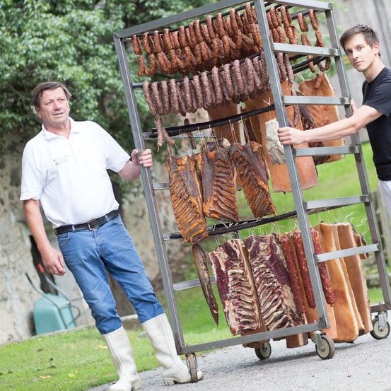Zwei Männer schieben einen Wagen mit verschiedenen Fleischsorten, einschließlich Würsten und Fleischstreifen, im Freien. Der Mann auf der linken Seite trägt weiße Stiefel und einen Gürtel.