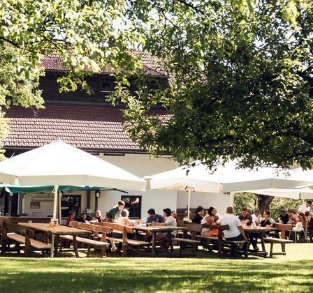 Eine Gruppe von Menschen isst im Freien in einem Restaurant unter großen weißen Sonnenschirmen, umgeben von Bäumen und Gras. Das Gebäude hat ein braunes Dach.