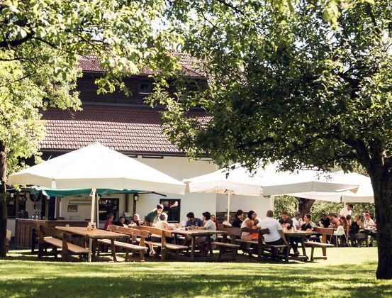 Eine Gruppe von Menschen isst im Freien in einem Restaurant unter großen weißen Sonnenschirmen, umgeben von Bäumen und Gras. Das Gebäude hat ein braunes Dach.