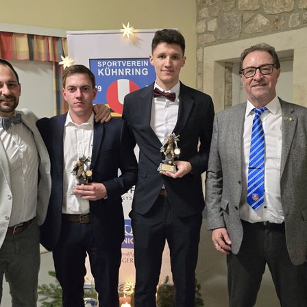 Vier Männer stehen lächelnd zusammen in formeller Kleidung, halten Trophäen, einer trägt eine Brille und eine Fliege, ein anderer einen Anzug mit Fliege und zwei in formellen Anzügen.