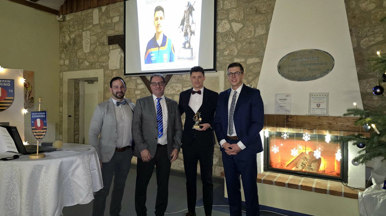 Vier Männer stehen vor einem Kamin mit einer Projektionsleinwand hinter ihnen, die ein Foto eines Mannes zeigt. Ein Mann hält einen Pokal.