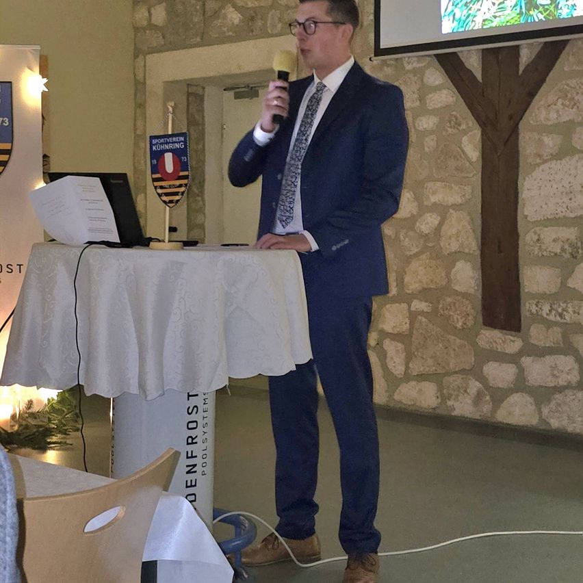 Ein Mann in einem blauen Anzug steht mit einem Mikrofon an einem Podium und spricht vor einem kleinen Publikum. Ein Tisch mit Laptop und Papieren steht vor ihm. Ein Banner mit Logo ist links angebracht, und eine Projektionsleinwand ist an der Wand montiert.