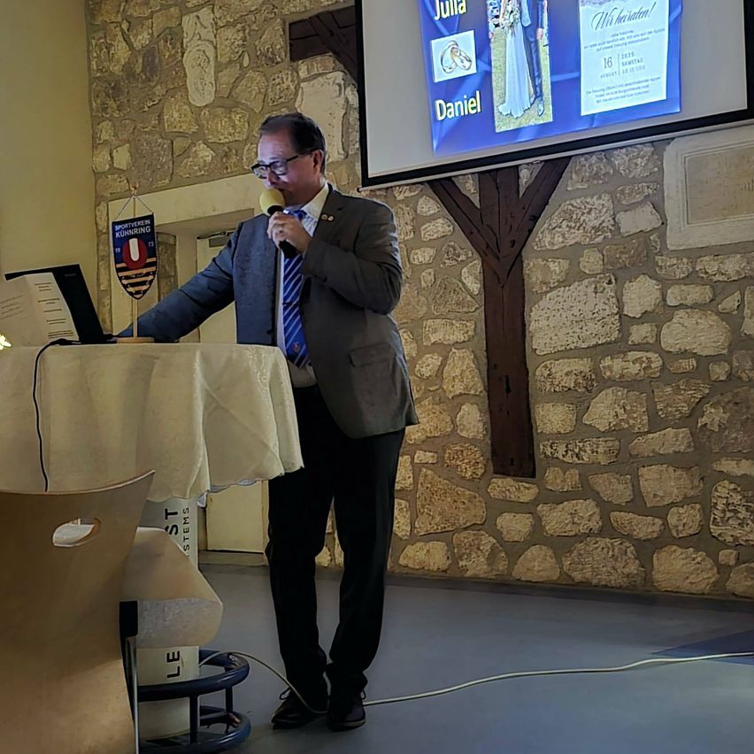 Ein Mann spricht in ein Mikrofon an einem Podium, wobei eine Leinwand hinter ihm Hochzeitsfotos zeigt.