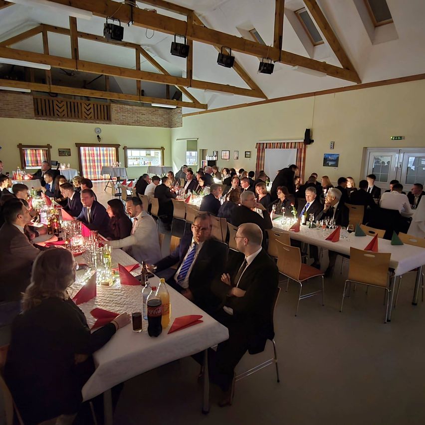 Eine große Gruppe von Menschen in Anzügen versammelt sich zu einer formellen Veranstaltung in einem Saal mit Holzbalken und Ziegelwänden. Lange Tische sind mit weißen Tischdecken bedeckt, und Getränke und Servietten sind ordentlich arrangiert.
