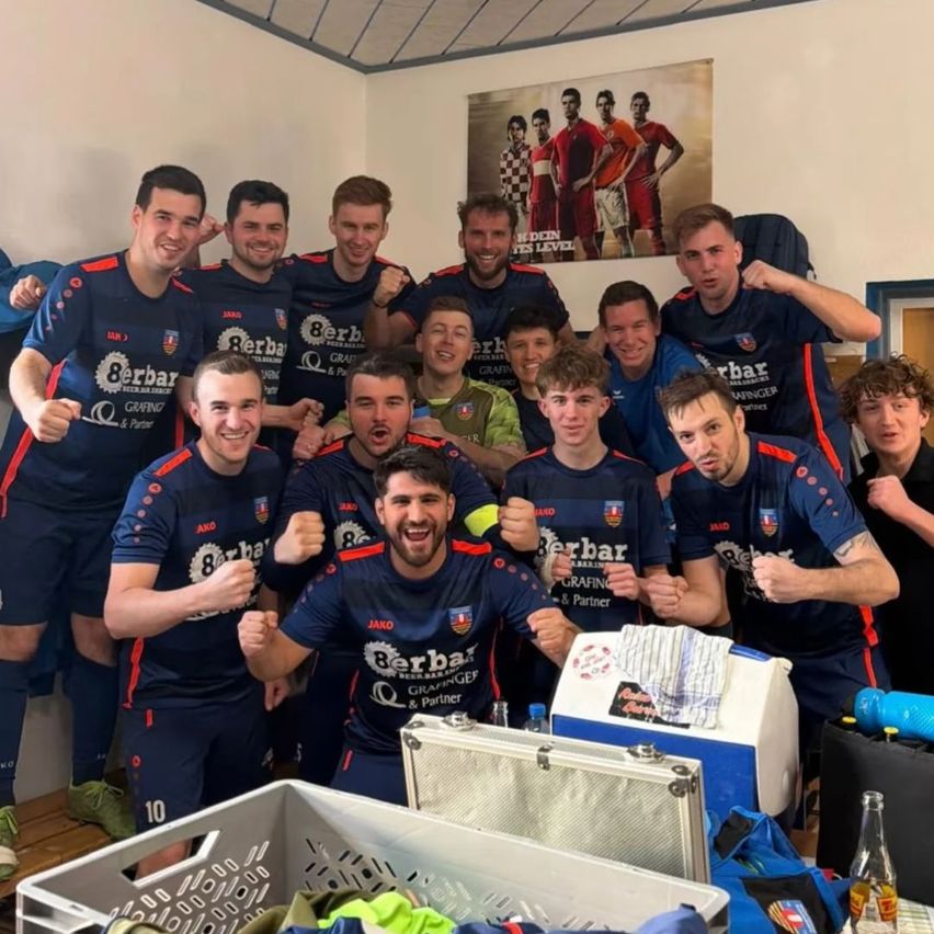 A soccer team in blue uniforms poses for a group photo in a locker room, with some players making fists and smiling. Equipment and bags are on the floor. A poster of players in red uniforms is on the wall.