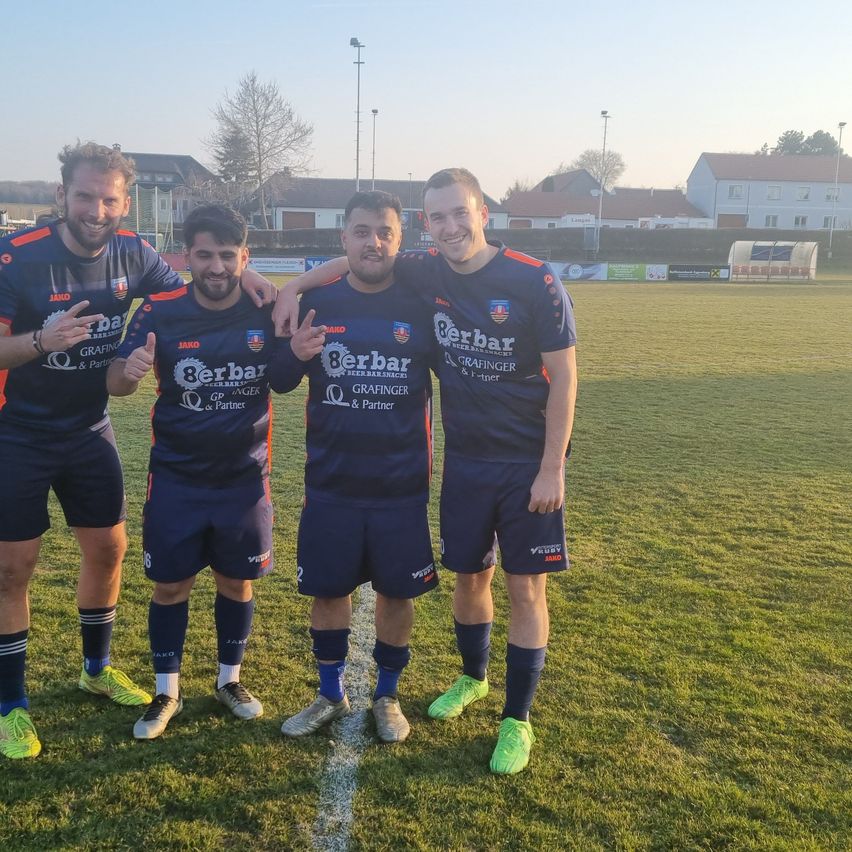 Four men in blue uniforms with various logos stand on a field, smiling, with a grassy background and houses in the distance.