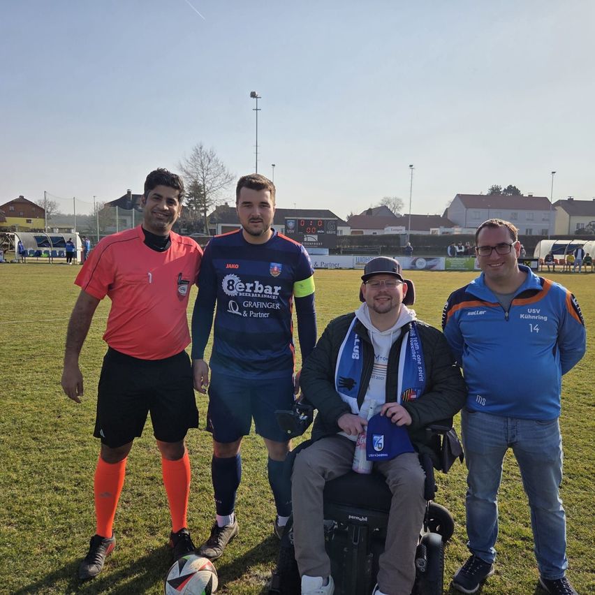 Four men stand on a soccer field. The man in the middle is in a wheelchair. Another holds a soccer ball. They are smiling and seem to be posing for a photo.