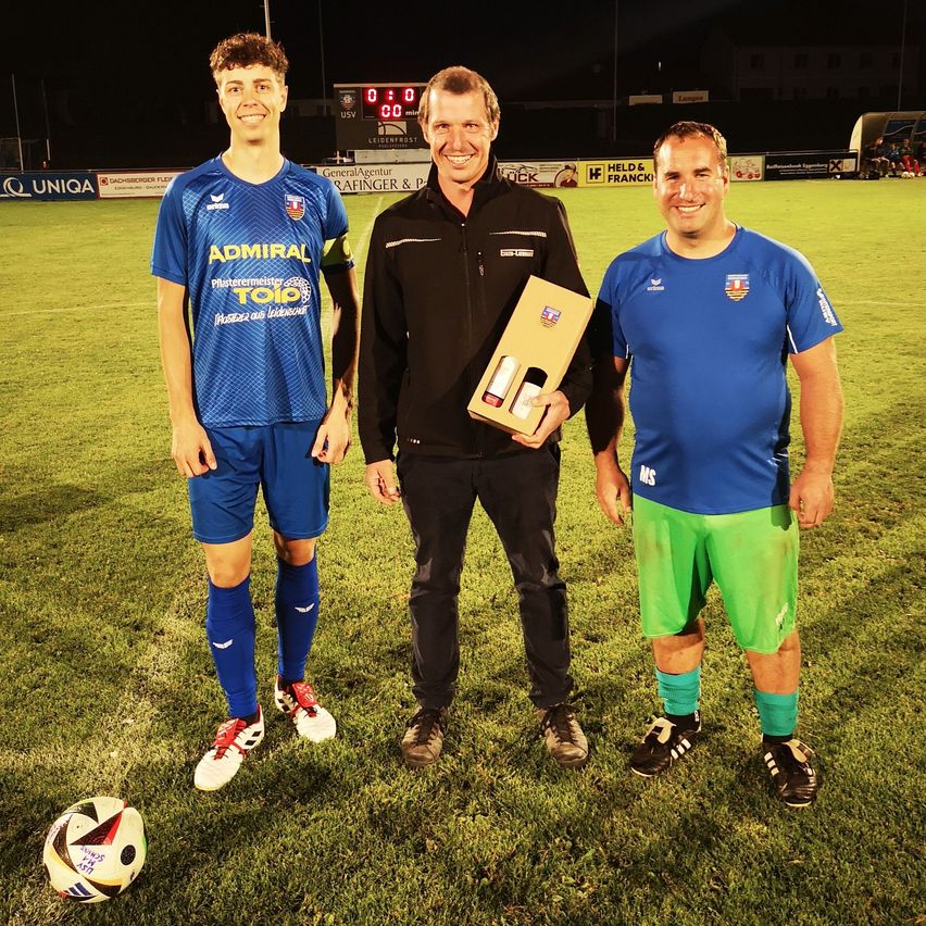 Drei Männer stehen auf einem Fußballfeld, zwei in blauen Trikots und einer in Schwarz, halten eine Box mit zwei Flaschen. Der Mann in der Mitte lächelt, und der Hintergrund zeigt eine Anzeigetafel und Werbung.