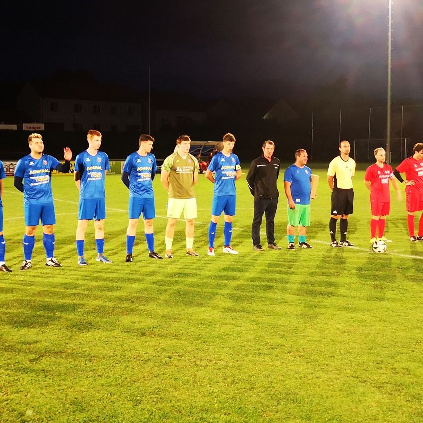 Eine Gruppe von Fußballspielern in blauen und roten Trikots steht bei Nacht auf einem Feld, mit einem Fußball auf dem Boden.