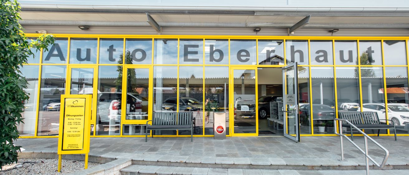 Ein Autohaus mit Glaswänden und gelben Rahmen. Bänke und ein Topf mit Pflanzen stehen draußen. Der Eingang ist offen und Autos sind drinnen geparkt.