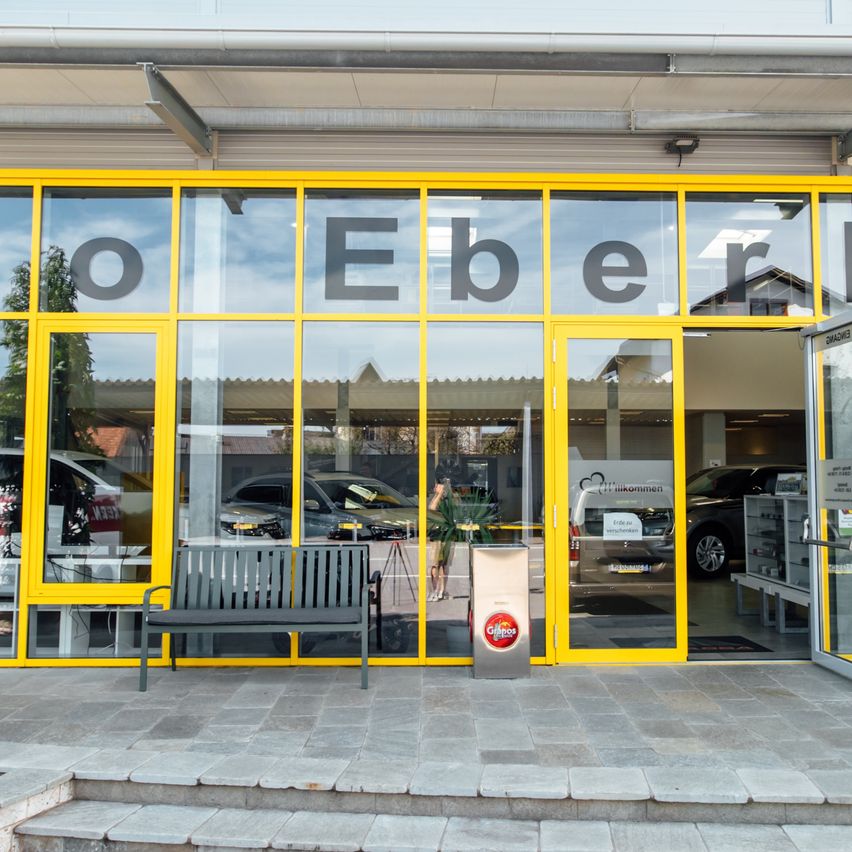 Ein Autohaus mit Glaswänden und gelben Rahmen. Bänke und ein Topf mit Pflanzen stehen draußen. Der Eingang ist offen und Autos sind drinnen geparkt.