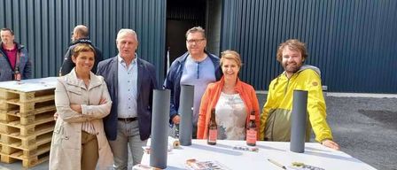 A group of people stand near a table with bottles and items in front of a building. A large metal structure is behind them.