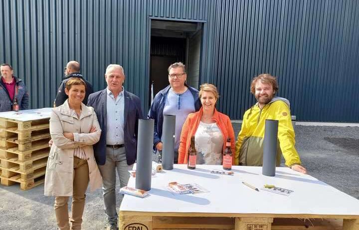 Eine Gruppe von Menschen steht neben einem Tisch mit Flaschen und Gegenständen vor einem Gebäude. Dahinter steht eine große Metallkonstruktion.
